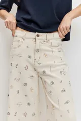 Pantalón de jean wide leg color beige con pequeños bordados y estampados coloridos dispersos, de tiro medio y corte amplio en las piernas.