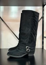 Bota alta de gamuza negra con caña alta, flecos laterales, tiras con hebillas metálicas en el tobillo y taco cuadrado.