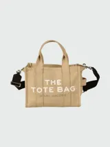 Bolso tipo tote de lona color beige de la marca Marc Jacobs. Presenta el texto 'THE TOTE BAG' estampado en el frente en color blanco, dos asas de mano superiores y una correa de hombro ajustable y removible en color negro.
