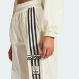 Pantalón deportivo Adidas de corte holgado en color blanco, con cintura elástica y diseño de tres tiras negras verticales en los laterales que incorporan detalles de red y el logo de la marca.