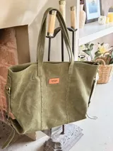 Bolso tipo tote bag de cuero gamuzado color verde militar, con asas de hombro y detalles de costuras decorativas. Cuenta con una etiqueta de cuero en el frente con la inscripción de la marca.