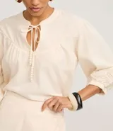 Blusa estilo bata color blanco roto, confeccionada en cambraia de algodón y lino. Presenta cuello con lazo ajustable, canesú fruncido en el busto y mangas tres cuartos con puños elásticos y bordados.