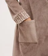 Bata de baño de plush color marrón, con mangas largas, cuello amplio, bolsillo delantero y lazo ajustable en la cintura. Presenta detalles de ribete estampado en los bordes y puños.