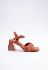 Sandalia de taco alto color tostado, fabricada en símil cuero, con tiras cruzadas en el empeine con pespuntes decorativos, tira con hebilla ajustable al tobillo y taco geométrico ancho con base transparente.