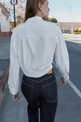 Camisa corta de cuello solapa y manga larga, con trabillas decorativas en los hombros. Presenta pliegues delanteros, elástico en la espalda y cierre frontal con botones.