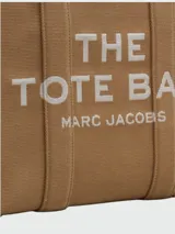 Bolso tipo tote de Marc Jacobs, modelo The Jacquard Large Tote Bag, confeccionado en tejido jacquard color tostado. Presenta el nombre de la marca y el modelo bordados en el frente en color blanco, doble asa superior y detalles de cintas laterales con inscripciones.