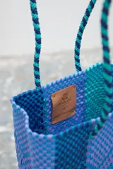 Bolso tipo matera tejido artesanalmente con tiras de plástico en tonos degradados de azul, violeta y turquesa, con diseño geométrico y correas trenzadas.