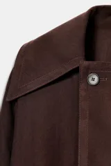 Chaqueta marrón oscuro con cuello solapa, manga larga ancha con trabilla y botón, y cinturón del mismo tejido anudado a la cintura. Cierre frontal cruzado con un botón visible.