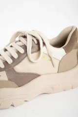 Zapatilla urbana de mujer con plataforma gruesa, combinando paneles de cuero en tonos beige, blanco roto y marrón topo. Presenta cordones anchos y un detalle de texto en el talón.