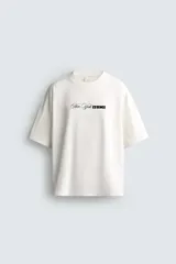 Camiseta de corte holgado con cuello redondo y manga corta. Presenta un diseño frontal con la frase New York Essence combinando tipografía cursiva y letras en bloque con efecto relieve.