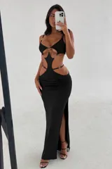 Vestido largo negro con diseño cut-out en la cintura y abdomen, decorado con argollas metálicas plateadas que conectan las piezas. Presenta breteles finos, escote pronunciado y una abertura lateral en la falda.