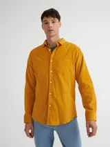 Camisa de manga larga color mostaza confeccionada en pana fina, con bolsillo en el pecho, botones de pasta, bordado en el ruedo y etiqueta de marca en la tapeta de los botones.