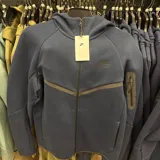 Campera deportiva Nike Tech Fleece color azul marino, con cierre frontal completo, capucha integrada y bolsillo vertical con cierre en la manga izquierda. Presenta diseño técnico con costuras selladas y logo de la marca en el pecho.