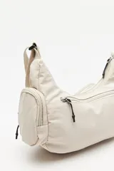 Cartera tipo shoulder color beige, de diseño utilitario y formato curvo. Confeccionada en material liviano y resistente, cuenta con correa regulable para llevar al hombro o cruzada. Presenta múltiples bolsillos con cierre en el frente y laterales, y un parche con el logo "Piece & Cake" en el frente.