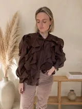 Camisa de color marrón chocolate, confeccionada en tela liviana y fluida. Presenta un diseño con múltiples vuelos frontales, cuello con lazo para anudar y mangas largas abullonadas con puños ajustados.