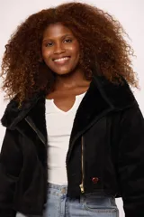 Campera corta tipo gamulán en color negro, con interior y detalles en puños y cuello de corderito sintético al tono. Presenta cierre frontal metálico y un botón decorativo en el ruedo.