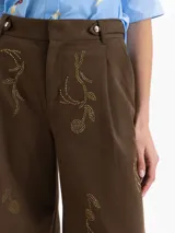 Pantalon de corte recto y tiro alto en color marron, con detalle de bordado en pedreria dorada sobre el frente. Presenta cierre frontal con botones y bolsillos laterales.