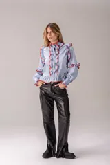Camisa celeste con volados decorativos en los hombros, pecho, mangas y ruedo, destacados con ribetes en tono rojo. Presenta cierre frontal de botones, cuello alto y mangas largas abullonadas con puños ajustados.