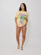 Remera color amarillo pastel con estampado de palmeras, flores de hibisco y texto 'California Ride the Tide Rusty Tribe'.