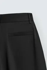 Pantalón negro de corte relaxed fit y estilo wide leg, con pliegues frontales dobles, bolsillos delanteros y detalle de bolsillos traseros de vivo. Cierre frontal cruzado con cremallera y gancho metálico oculto.
