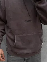Hoodie de color marrón con efecto acid wash, capucha con cordones ajustables, bolsillo canguro frontal y pequeño estampado con texto en el pecho. Cuenta con una etiqueta decorativa en el borde inferior.