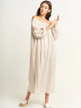 Vestido largo color beige, con escote elastizado que permite usarlo con los hombros descubiertos, mangas largas abullonadas, bolsillos laterales y tajos laterales en el ruedo.