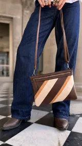 Sobre de mano con diseño de franjas diagonales en tonos marrones y beige, confeccionado en símil cuero. Cuenta con cierre metálico superior, correa desmontable para usar como bandolera, bolsillo interno con cierre y compartimento abierto.