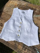 Chaleco de lino en color beige, con escote en V, cierre frontal con botones y bolsillos delanteros decorativos.