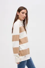 Remera tipo polo de manga larga con diseño de rayas horizontales anchas en bloques de color crema y beige. Presenta cuello camisero con cierre de botones y un pequeño bordado en el pecho.