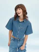 Camisa corta de denim azul claro, con cuello camisero, manga corta ancha y cierre frontal de botones blancos. Presenta un bolsillo de parche en el pecho izquierdo.