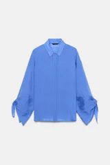 Camisa de cuello solapa y manga larga con detalle de lazos en los puños. Presenta un diseño de rayas verticales a tono y cierre frontal con botones ocultos.