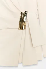 Blazer de corte estructurado en color beige, con cuello solapa y manga larga. Presenta un diseño cruzado con cierre mediante lazada del mismo tejido y un distintivo aplique metálico dorado en el frente.