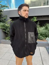 Campera de abrigo negra de tela peluda con cierre frontal, cuello alto, bolsillos laterales y un bolsillo impermeable en el pecho.