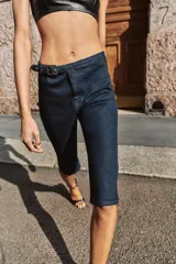 Jeans de tiro medio en denim azul oscuro, con diseño capri de pernera entallada. Presenta cierre frontal con cremallera y botón, y un cinturón integrado con hebilla metálica en la cintura.