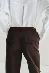 Pantalón chino de tiro bajo color marrón oscuro, con pernera recta y bajo con vuelta. Incluye un cinturón trenzado delgado de color marrón claro. Cuenta con bolsillos delanteros y cierre frontal con cremallera y botón.