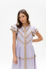 Vestido largo de lino color lila, con volados en las mangas y detalles de ribete en color beige que forman patrones en zigzag en el torso y el ruedo. Presenta un frente abotonado con una tira central beige.
