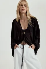 Top de crepe blanco con estampado de lunares negros, escote en V y cierre frontal con finas lazadas. Presenta mangas largas abullonadas con puños elásticos y bajo redondeado.