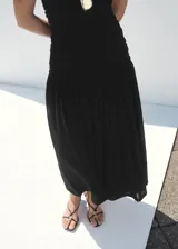 Vestido midi negro sin mangas, con cuello redondo y diseño ajustado en el torso mediante fruncidos laterales, que se abre en una falda evasé fluida.