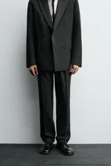 Pantalón de vestir negro con pliegues frontales, corte regular fit, bolsillos laterales y traseros de doble vivo con botón. Cierre frontal cruzado con cremallera y gancho metálico.