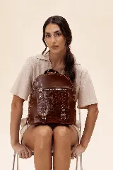 Mochila de cuero vacuno color marrón, con diseño estructurado y combinación de textura croco en el bolsillo frontal. Cuenta con un compartimento principal con cierre, un bolsillo frontal con cierre metálico y hebillas decorativas, y correas regulables.