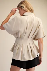 Camisa de popelina color beige con diseño estructurado, presenta pliegues verticales en la cintura que crean un efecto entallado y mangas cortas con volumen. Cuenta con cuello clásico y cierre frontal de botones.