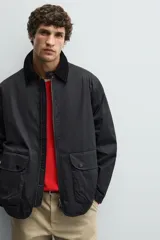 Parka de corte relajado en color negro, confeccionada en tejido de algodón con interior ligeramente acolchado. Presenta cuello de solapa en pana, cierre frontal con cremallera y bolsillos delanteros tipo plastrón con solapa y botones a presión.
