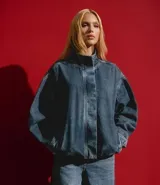 Campera bomber de jean azul con cuello alto estructurado, cierre frontal oculto, mangas largas con puños ajustados y ruedo con efecto fruncido. Cuenta con bolsillos embutidos y diseño de corte holgado.