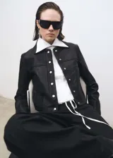 Chaqueta de cuero ovino en color negro con acabado en napa. Presenta cuello camisero, cierre frontal con botones a presión metálicos, bolsillos laterales y puños abotonados.