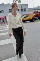 Blusa de color beige con escote en pico y lazo frontal decorado con abalorios. Presenta mangas largas con puños elásticos y un detalle de volantes amplios con terminación de puntilla a tono sobre los hombros y el pecho.