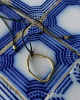 Collar con dije colgante de forma orgánica e irregular, acabado dorado, suspendido de un cordón negro de algodón natural.