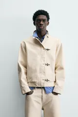 Cazadora de algodón color beige claro, corte relajado, con cuello subido y manga larga. Cierre frontal con cremallera oculta por solapa con cierres metálicos tipo alamar.