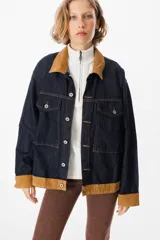 Chaqueta de denim azul oscuro con cuello y puños en pana color tostado. Presenta cierre frontal con botones metálicos y dos bolsillos frontales con solapa.
