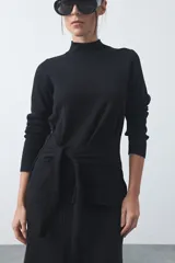 Polera de tejido de punto color beige, con cuello alto, manga larga y detalle de lazo ajustable en la cintura que aporta estructura a la silueta.