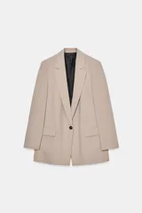 Blazer de corte recto en color beige, con cuello solapa, manga larga y cierre frontal con un solo botón. Presenta bolsillos delanteros con solapa y forro interior en color negro.